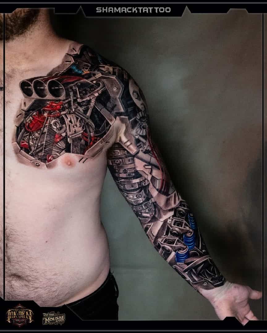 Person showing an intricate black, red, and blue biomechanical tattoo sleeve and chest piece against a gray wall